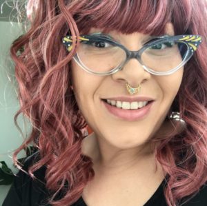 A light brown skinned mixed Black femme smiles at the camera. They have mauve hair, blue and gold cat eye glasses, and a gold and turquoise septum ring.