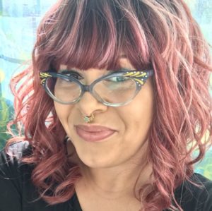 A light brown skinned mixed Black femme smiles at the camera. They have mauve hair, blue and gold cat eye glasses, and a gold and turquoise septum ring.