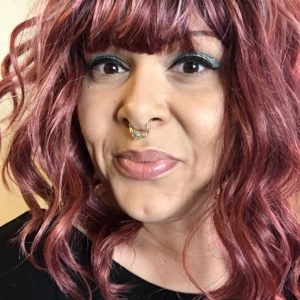 A light brown skinned mixed Black femme smiles mischievously at the camera. They have mauve hair and are wearing sparkly turquoise eyeshadow, black eyeliner, and a rose gold and turquoise septum ring.